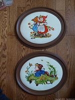 Top view of two oval wood framed stitched textile artworks on wooden floor, each showing children in vintage style.