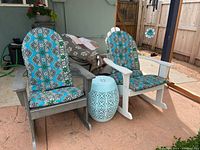 Two Adirondack rocking chairs (one grey, one white) with turquoise patterned cushions and a light turquoise metal side table between them