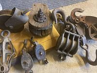 Top view of various pulleys including wooden wheel mounted on metal frames, metal pulleys with hooks and eyes, displayed on a cardboard surface on the floor.