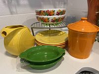 Collection of nine Fiestaware ceramic pieces arranged on countertop including covered jar, pitcher, plates, oval dishes, and two large serving bowls with leaf motif.