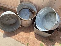 Five galvanized metal tubs, four round and one square, some showing rust, dirt and wear.