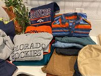 Folded men's clothing including Denver Broncos and Colorado Rockies sweatshirts, striped button-up shirt, and pants