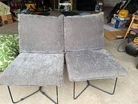 Pair of gray upholstered indoor lounge chairs with black metal legs placed side by side in a room with boxes and equipment.