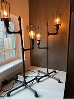 Pair of industrial metal table lamps illuminated with Edison bulbs and wire cage covers, standing on a white surface near a window and wall.