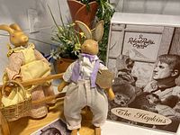 Photo showing two ceramic bunny figurines, one female holding a basket, one male dressed in white pants and lavender shirt, alongside original decorative box labeled 'Robert Raikes Originals The Hopkins'.