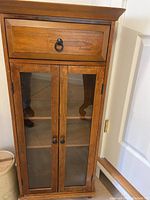 Side view of the wooden side table showing one drawer and two glass doors with black knobs.