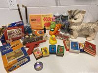 Overview photo showing assortment of vintage games, puzzles, toys, and figurines including a large ceramic cat, plastic toy gun, rubber duck, and boxed puzzles and games on a kitchen counter.