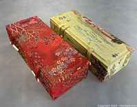 Two rectangular fabric-covered boxes, one red with floral gold designs and one yellow-green with scenic calligraphy patterns, displayed side by side on a gray background to show exterior appearance and size comparison.