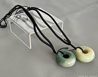 Photo of two jade donut-shaped pendants on black cords, displayed on a clear acrylic stand, showing the color and texture of both pendants.