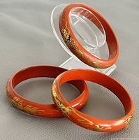 Three Chinese red lacquer bangles with gold and black dragon hand painting displayed on a clear stand.