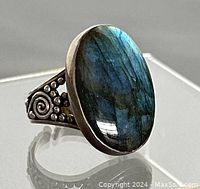 Side angled front view showing oval labradorite gemstone set in silver bezel with visible blue-green iris color shift and decorative band with small spiral and dot embossing.