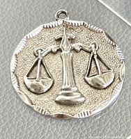 Close-up front view of the round silver medallion pendant showing raised Libra scales on a textured background with scalloped edge