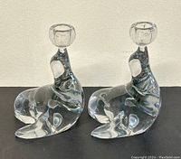 Pair of clear art glass candle holders in the shape of seals, front-side view on dark table against light wall background.