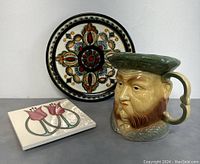 Front view of pottery plate with colorful detailed pattern, Henry VIII Toby mug with handle, and decorative tile with tulips.