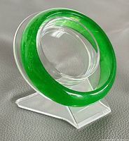 Natural green jade bangle shown on clear acrylic display stand, highlighting rich green translucent color and smooth polished surface with minor wear.