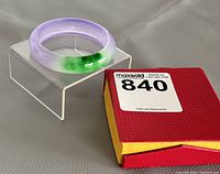Photo of multicolor jade bangle on clear stand beside original red and yellow box with lot label.
