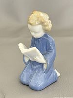 Front angle of the porcelain figurine showing the girl reading a book kneeling, with detailed view of hair and dress.
