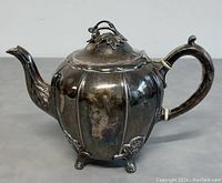 Front and side view of vintage silver plated teapot showing scalloped body, floral foot decorations, curved handle with white spacer rings, and spout with detailed tip.