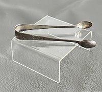 Full view of the vintage sterling silver sugar tongs displayed on a clear plastic stand, showing the shape and design of the tongs.
