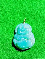Front view of light green natural jade carved Buddha pendant with 14k gold bail highlighted by bright lighting on a green textured surface.
