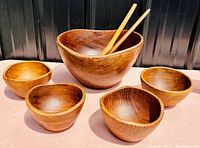 Wide view of the teak salad bowl set including one large bowl, four smaller bowls, two serving utensils on a pink surface against a black background.