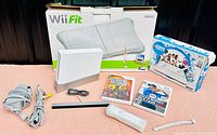 Overall display of Nintendo Wii console, Wii Fit balance board box, two Wii game cases, Wii Remote, and sensor bar on pink carpet.