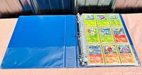 Blue binder open showing a page with nine Pokemon trading cards in sleeve pockets arranged 3x3.