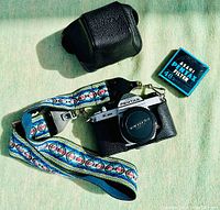 Top view of Asahi Pentax K1000 film camera with colorful strap, black leather camera case, and boxed Asahi Pentax 46mm filter.