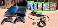 Xbox One console, two controllers, power adapter with cords, and five game cases displayed on a flat surface under daylight.