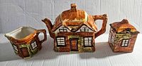 Front view of teapot, creamer, sugar container shaped as rustic cottages with brown and beige glaze.