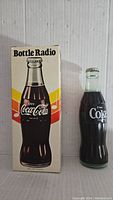 Photo of two vintage Coca-Cola bottle-shaped transistor radios, one displayed with original box. The radios resemble classic ribbed glass Coca-Cola bottles with branded labels and bottle cap tops.