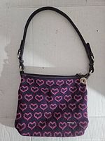 Full front view of purple crochet purse with pink hearts and black strap, placed against white background.