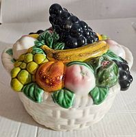 Close-up angled view of the top of the ceramic fruit basket lid showing fruits such as blackberries, banana, grapes, peaches and leaves in detailed, colorful 3D design.