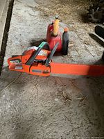 Overview of chainsaw on concrete floor with red plastic fuel can and black oil bottle behind