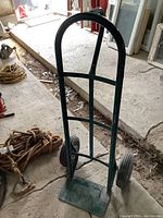 Front view of green metal dolly standing upright, showing full height and base plate with wheels on a concrete floor.