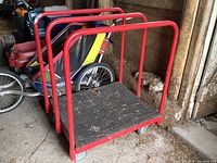 Front and side perspective of metal flat dolly showing red frame, black platform, and wheels on ground dusted with debris.