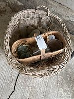 Large wicker basket containing a wooden fruit basket, glass bottles, canning jar lids, and one jar, on concrete floor.
