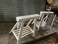 Two white wooden IKEA Finnvard trestles and one white foldable chair resting on a concrete surface in front of a corrugated metal door. The trestles have slatted lower shelves and show dirt and wear.