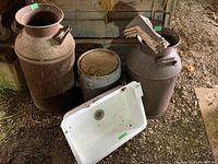 Two large rusted metal milk cans next to white enamel sink and other items on ground in barn