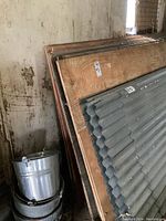 Stack of plywood sheets leaning against wall alongside corrugated metal panel and metal buckets