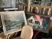 Two framed vintage prints leaning on a metal chair and a rustic wooden railing; one print shows signs of wear and water damage