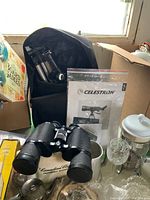 Celestron telescope manual and tripod partially visible behind Tasco binoculars on table with other items around.