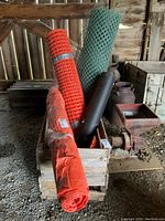 Photo of two fencing rolls (green and orange mesh), black tar paper roll, and orange plastic tarp roll placed inside a wooden crate in a barn setting.