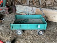 Green metal utility wagon with sides raised, positioned in barn, showing dirt ground and surrounding farm equipment.