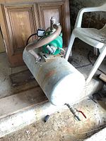 Side view of green electric water pump and motor mounted on a rusty horizontal metal tank with hose connected at the top and placed next to a white plastic chair.