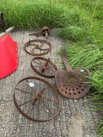 Lot of rusted old metal parts including three spoked tractor wheels laid out on ground with some grass around.