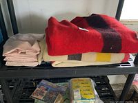 Overview of three folded wool blankets with stripes and stack of six pale pink pillowcases on black shelving