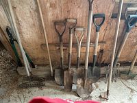 Photo showing 4 shovels with D-grip wooden handles, 2 spades, 2 garden forks and 2 metal rakes, all with wooden handles, standing against a wood backdrop on concrete floor.