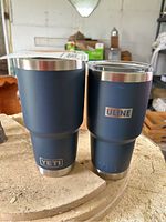 Two blue Yeti travel mugs, one with Yeti logo, one with Uline logo, placed on a rough round surface.