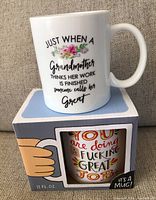 Photo shows one white ceramic mug with the quote about grandmother and flowers, sitting on a box. The box displays part of a second mug with a colorful motivational quote.